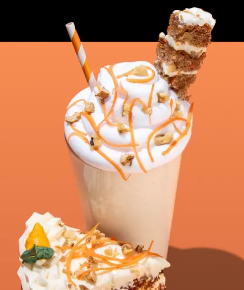 A tall vanilla shake topped with a large swirl of whipped cream, orange drizzle, and chopped walnuts. A piece of carrot cake is skewered on top alongside an orange and white striped straw. A slice of carrot cake with white frosting and an icing carrot decoration is in the foreground against an orange background.