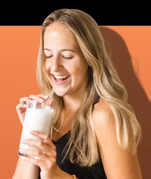 A smiling woman with long blonde hair holds a clear glass of a white shake and drinks from a straw. She is wearing a black top and stands against a solid orange background.