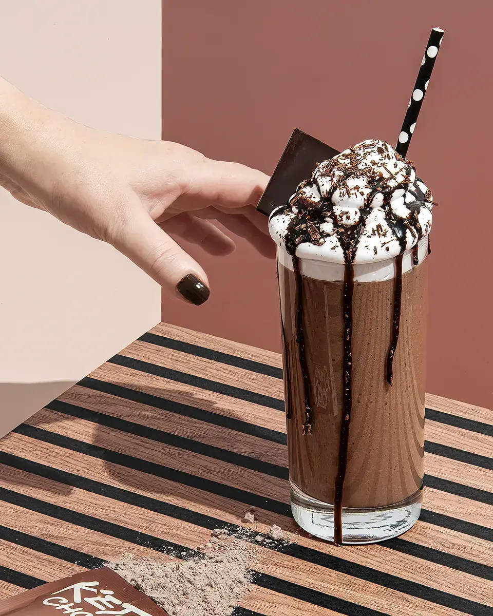 A colorful product selection grid against a blue background. The grid displays individual dessert items, each in its own box. Some boxes are in full color, like a croissant pastry, a coffee drink, and a slice of pie with berries. Other boxes are in black and white. A yellow circle with x1 is in the upper right corner of some boxes. A hand icon with a cursor points to a yellow and white box containing a peeled banana.