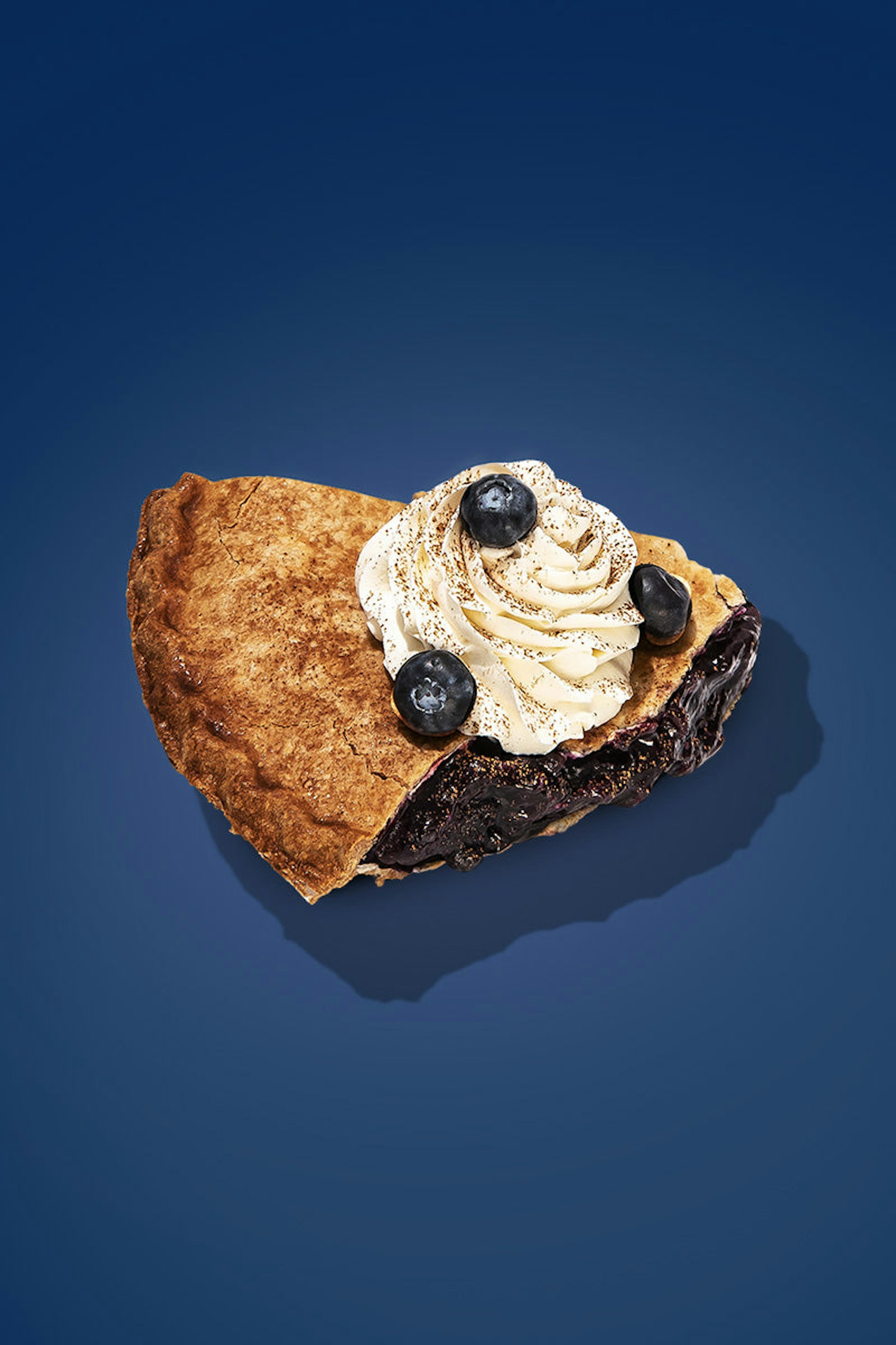 A slice of double-crusted blueberry pie topped with whipped cream and more blueberries