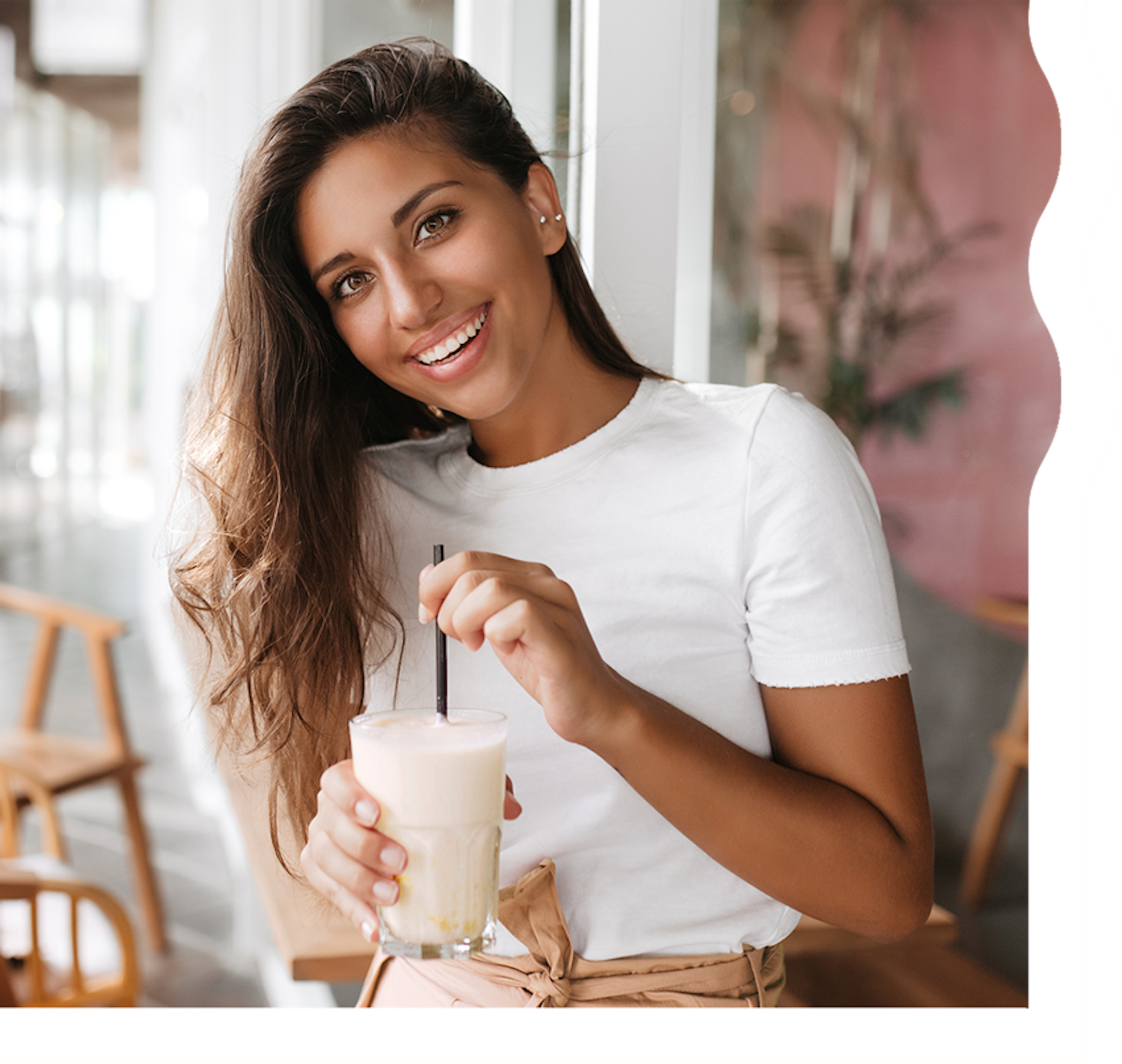 A woman with tan skin and long, dark brown hair is smiling and looking directly at the camera. She is holding a white, creamy shake.