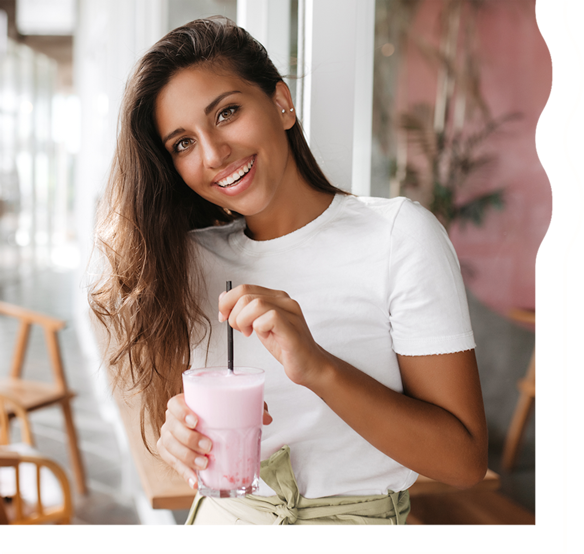 A woman with tan skin and long, dark brown hair is smiling and looking directly at the camera. She is holding a delicately pinkish red shake.