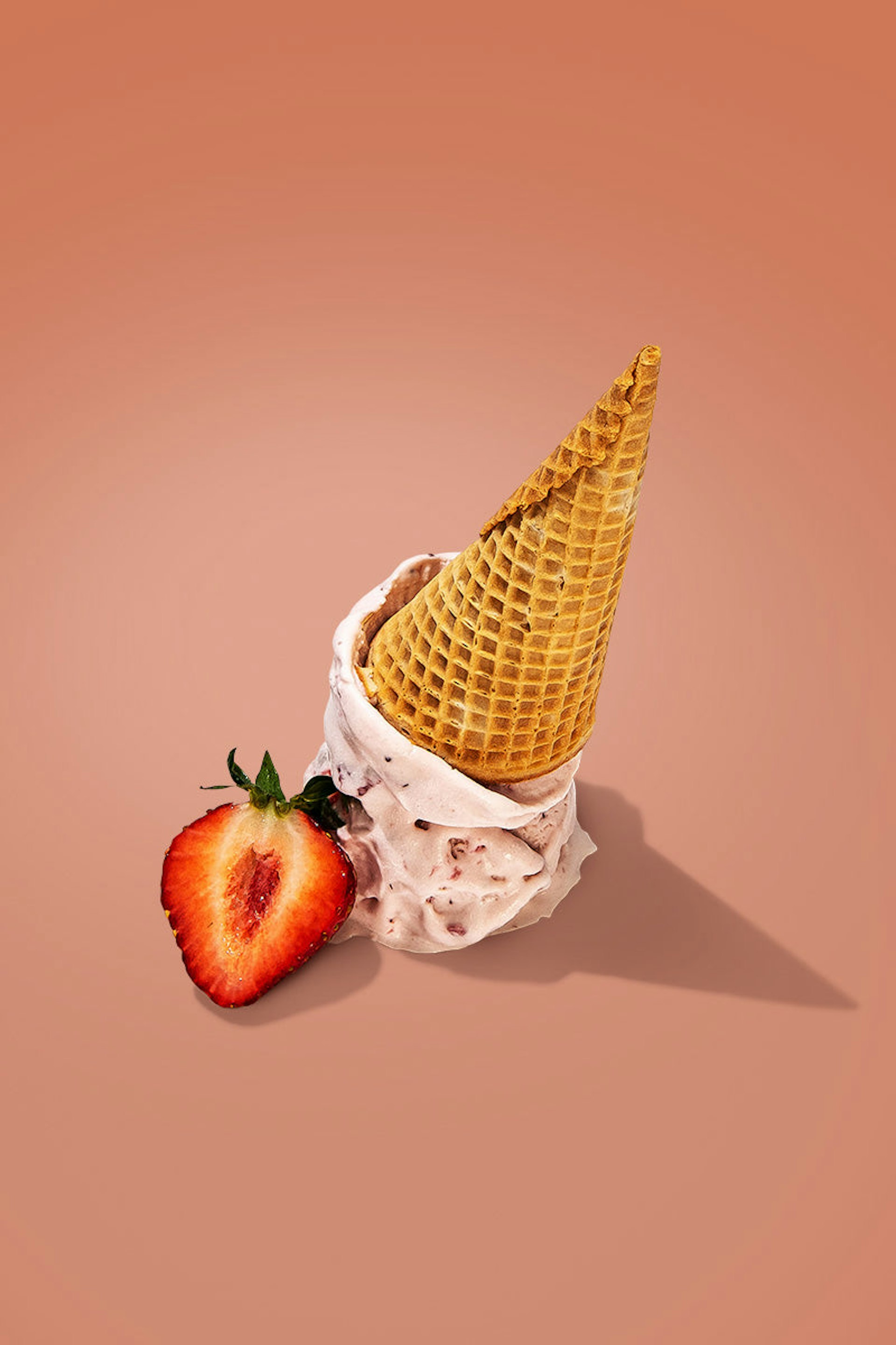 An upside ice cream cone on top of a few scoops of strawberry ice cream, with a sliced strawberry to the side