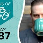 Digital banner featuring 100 DAYS OF KETO CHOW in white text with a cup and splash logo on a teal hexagon. Below it, DAY 087 is in black text on a light blue honeycomb pattern. A man drinks from a teal ribbed mug on the right against a wooden background.