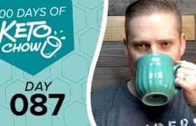 Digital banner featuring 100 DAYS OF KETO CHOW in white text with a cup and splash logo on a teal hexagon. Below it, DAY 087 is in black text on a light blue honeycomb pattern. A man drinks from a teal ribbed mug on the right against a wooden background.