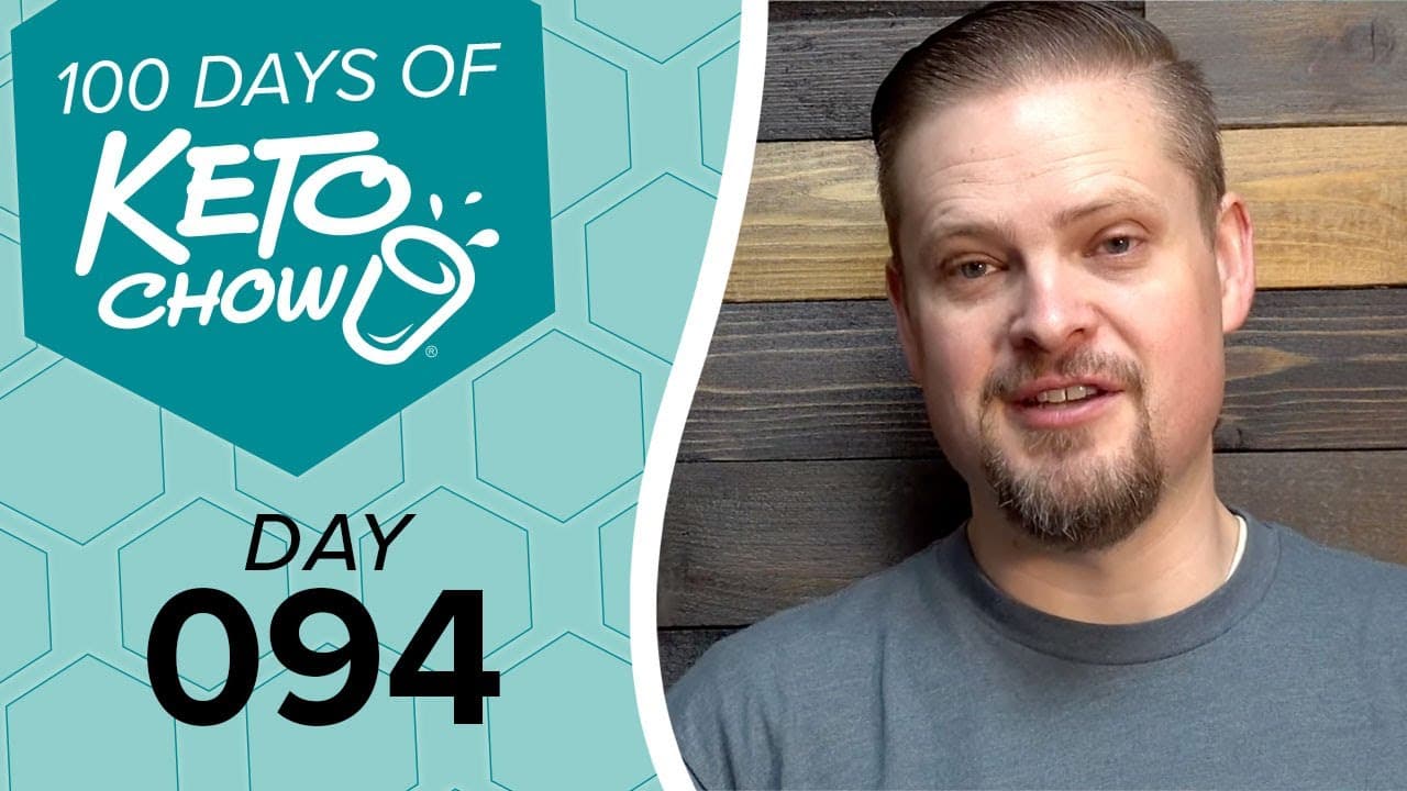 Promotional image featuring KETO CHOW branding. A teal graphic with a hexagonal pattern displays 100 DAYS OF KETO CHOW logo and text DAY 094. A man in a grey shirt smiles on the right against a dark wood background.