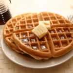 Three golden waffles with butter and syrup on a white plate, a syrup dispenser, and cutlery on a light brown woven placemat.