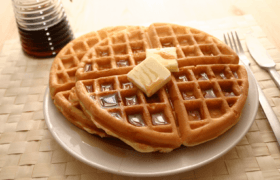 Three golden waffles with butter and syrup on a white plate, a syrup dispenser, and cutlery on a light brown woven placemat.