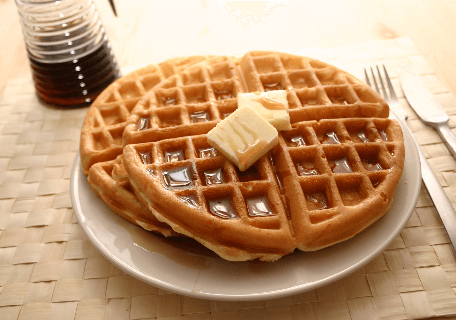 Three golden waffles with butter and syrup on a white plate, a syrup dispenser, and cutlery on a light brown woven placemat.