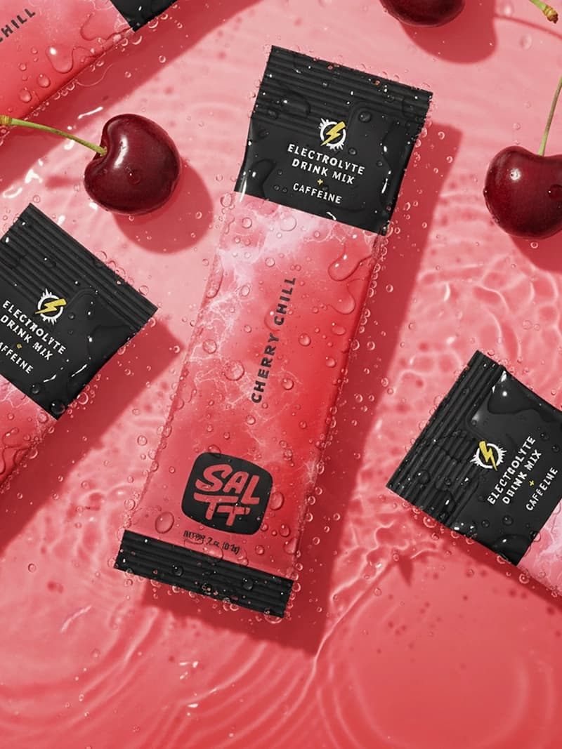 Packets of cherry-flavored electrolyte drink mix with caffeine, scattered on a wet pink surface with fresh cherries, conveying a fresh, energizing vibe.
