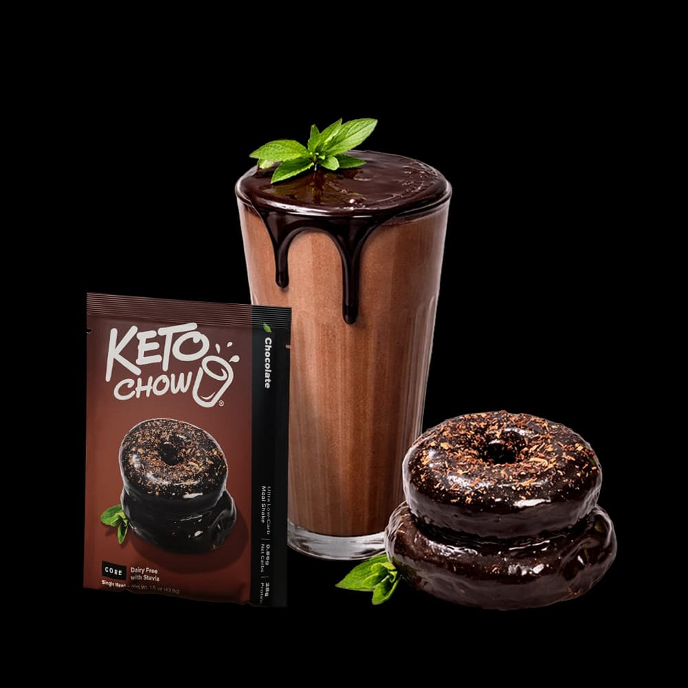A single-serve Keto Chow Chocolate packet stands next to a tall glass of chocolate shake topped with chocolate syrup and a mint sprig. Two chocolate-glazed donuts are stacked in the foreground. The entire composition is set against a solid black background.