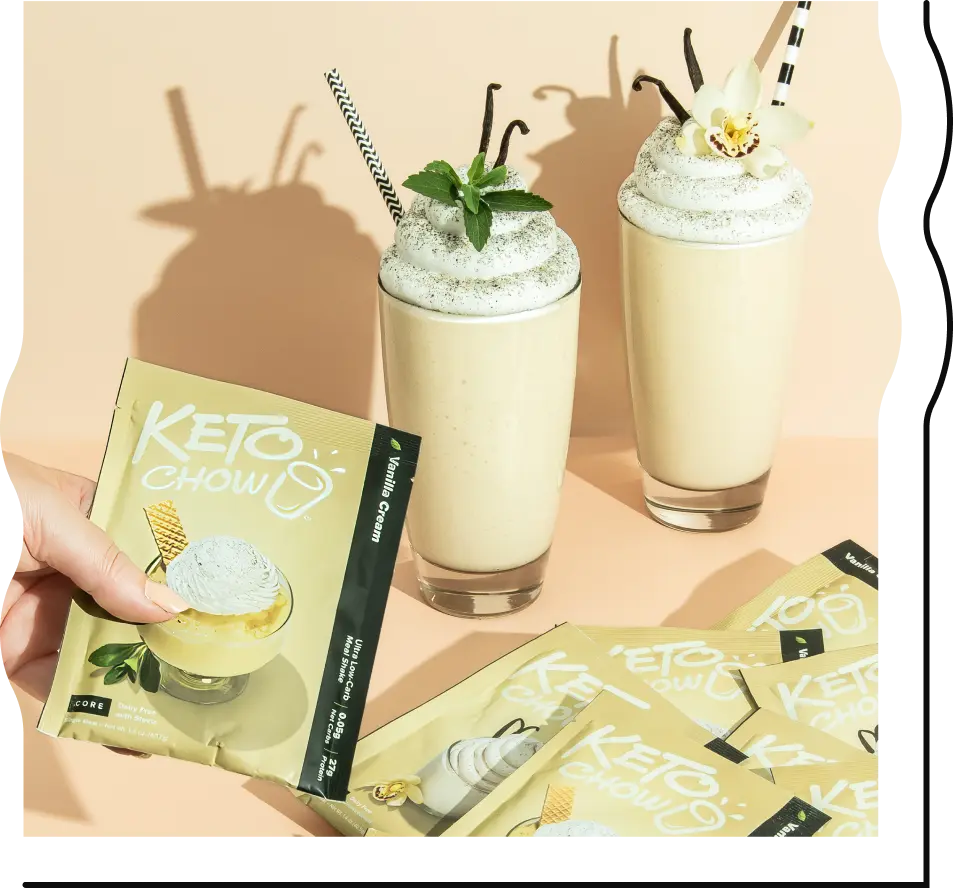 A hand holds a Keto Chow Vanilla Cream single-serve packet in front of two tall glasses of vanilla shakes. The shakes are topped with whipped cream, vanilla beans, mint leaves, and a striped straw. Several more Keto Chow packets are scattered in the foreground on a pale peach background.