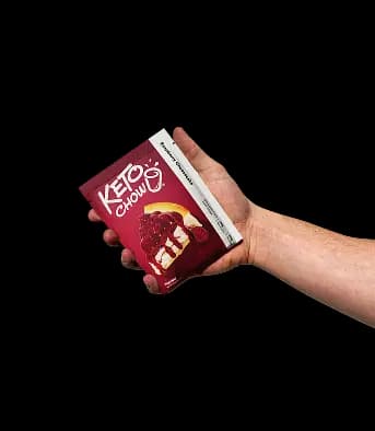 A hand holds a single-serve Keto Chow Raspberry Cheesecake packet with a soft shadow. The dark red packet features the Keto Chow logo and a graphic of a slice of cheesecake topped with raspberries.