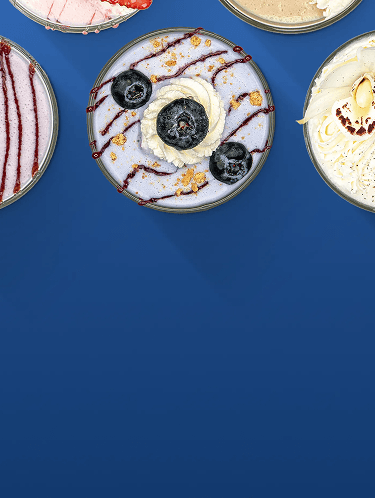 Four circular dessert bowls are on a solid blue background. A pink dessert with strawberries is at the top. A tan dessert with cookies is at the top right. A light purple dessert with blueberries, whipped cream, and chocolate syrup is at the center. A yellow dessert with a flower is at the right.
