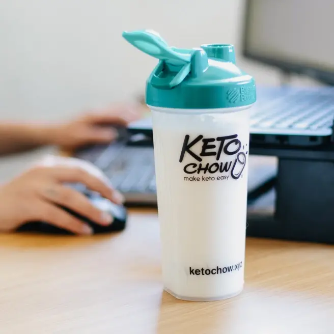 A Keto Chow shaker bottle with a teal lid sits on a wooden desk. In the blurred background, a person is using a computer keyboard and mouse. The bottle features the Keto Chow logo and the website ketochow.xyz.