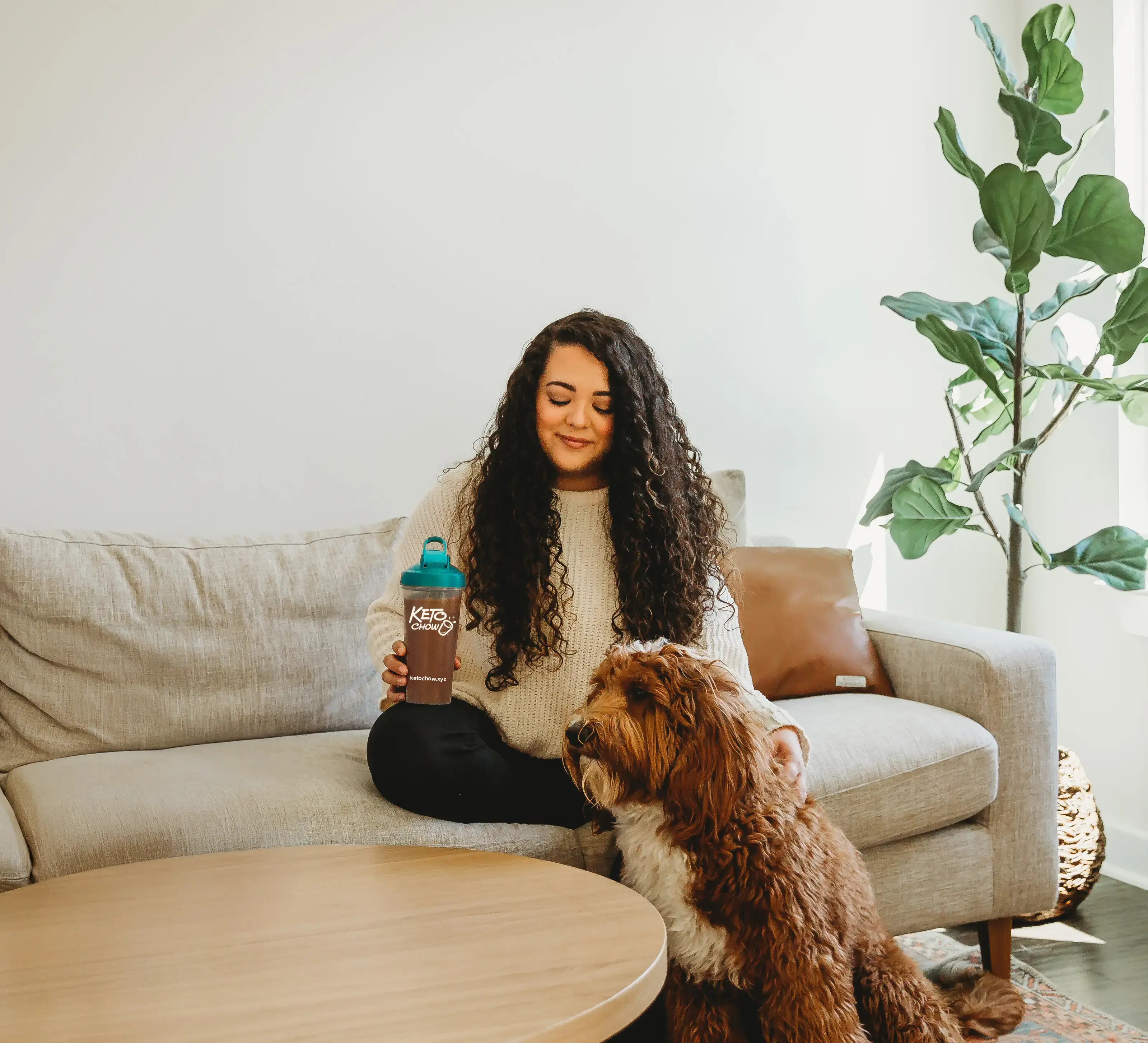 A woman with long curly hair sits on a light gray sofa holding a Keto Chow shaker bottle with a teal lid. A large brown dog sits next to her. The living room setting includes a wooden coffee table, a brown pillow, and a fiddle leaf fig plant in the background.