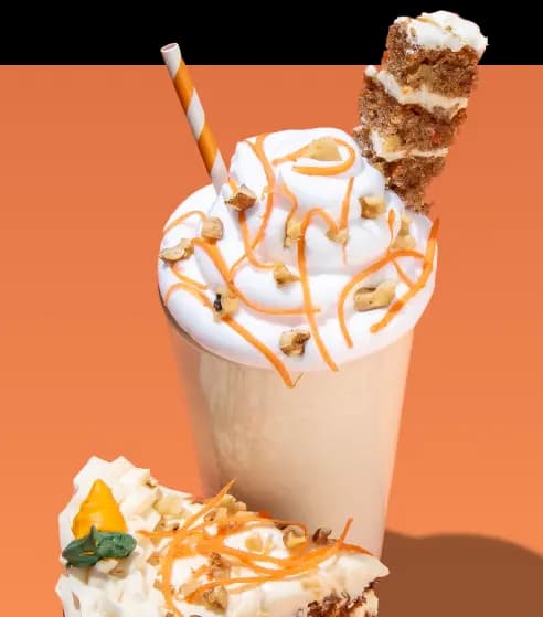 A tall vanilla shake topped with a large swirl of whipped cream, orange drizzle, and chopped walnuts. A piece of carrot cake is skewered on top alongside an orange and white striped straw. A slice of carrot cake with white frosting and an icing carrot decoration is in the foreground against an orange background.
