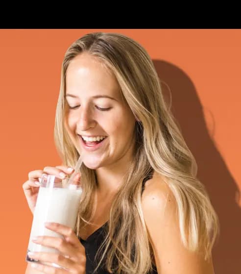 A smiling woman with long blonde hair holds a clear glass of a white shake and drinks from a straw. She is wearing a black top and stands against a solid orange background.