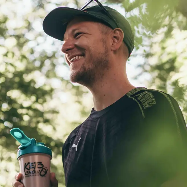 A man wearing a green baseball cap and black athletic shirt smiles while holding a Keto Chow shaker bottle with a teal lid. The background is a soft-focus view of green trees in natural sunlight.