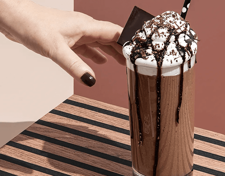 A hand reaches for a tall glass of chocolate shake topped with whipped cream, chocolate syrup, and chocolate shavings.