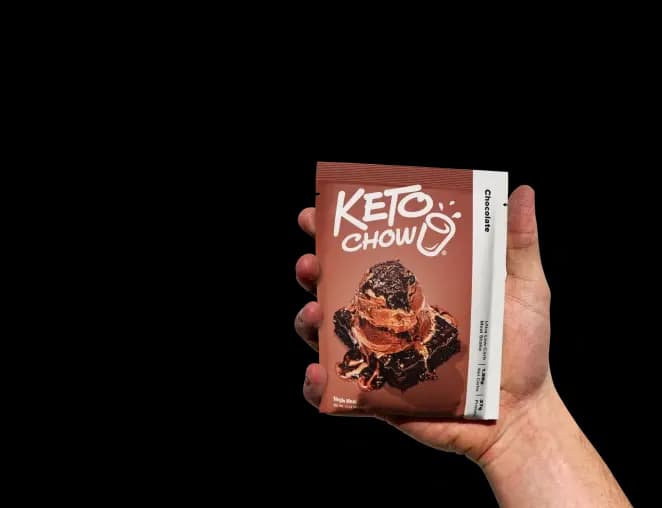 A hand holds a single-serve Keto Chow Chocolate packet with a soft shadow. The brown packet features the Keto Chow logo and a graphic of a chocolate brownie topped with a scoop of chocolate ice cream.