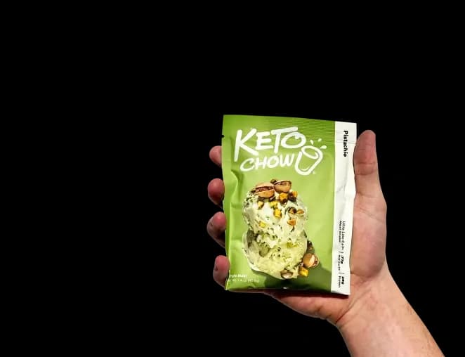 A hand holds a single-serve Keto Chow Pistachio packet with a soft shadow. The light green packet features the Keto Chow logo and a graphic of two scoops of pistachio ice cream topped with whole and crushed pistachios.
