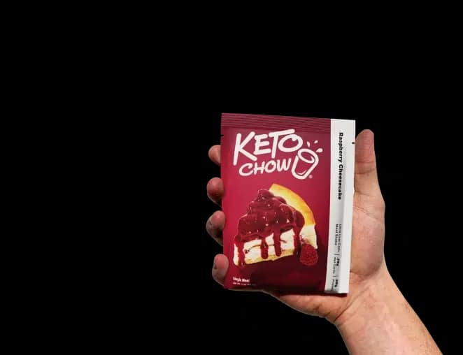 A hand holds a single-serve Keto Chow Raspberry Cheesecake packet with a soft shadow. The dark red packet features the Keto Chow logo and a graphic of a slice of cheesecake topped with raspberries.