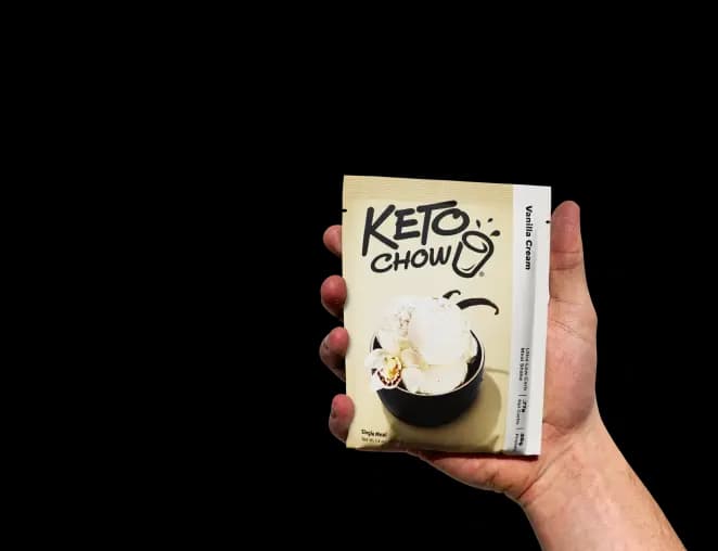 A hand holds a single-serve Keto Chow Vanilla Cream packet with a soft shadow. The cream-colored packet features the Keto Chow logo and a graphic of a white orchid flower and vanilla beans.