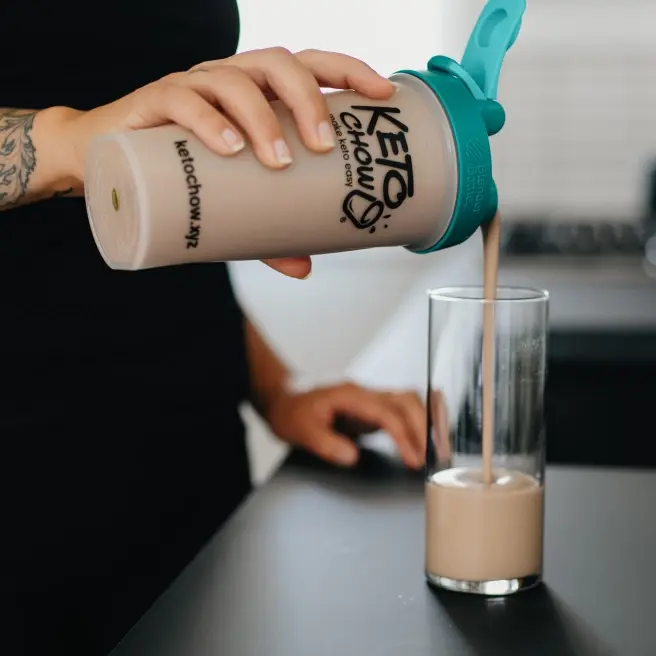 A person pours a chocolate-colored shake from a Keto Chow BlenderBottle with a teal lid into a clear glass. The shaker bottle features the Keto Chow logo and ketochow.xyz web address. The scene is set against a dark countertop and a neutral background.