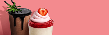 Two beverages are on a pink background. On the left is a chocolate shake with chocolate syrup dripping down the side and a sprig of mint on top. On the right is a layered dessert in a glass, with a white base, a red layer, pink whipped cream, and a strawberry on top.