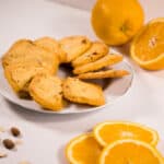 A white plate arranged with Orange Almond Shortbread cookies, surrounded by sliced oranges and almonds.