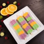 Pink, orange and green popsicles on a white tray surrounded by raspberries and sliced oranges and limes.