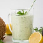 Close up-glass mug with green smoothie. Green and white straw in the smoothie. Surrounded by lemons, ginger and a coconut.