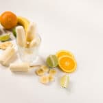 Three Banana Daquiri popsicles in a glass jar with two more laying to the side. Slices of bananas, oranges and limes surround the jar.