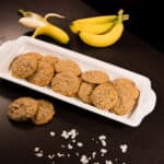 White tray filled with banana oatmeal cookies on a dark table. Bananas, coconut, and more cookies surround the plate.