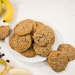 Banana oatmeal cookies mounded on a white plate, surrounded by bananas and sugar free chocolate chips.