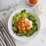 Close up of white bowl with sliced cucumbers, celery, green onions, and shredded buffalo chicken. A cup of cocktail sauce on the side