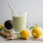 Distant view-Close up-glass mug with green smoothie. Green and white straw in the smoothie. Surrounded by lemons, ginger and a coconut.