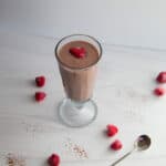 Single, tall glass with smoothie, topped with raspberries. Raspberries on table around the glass.