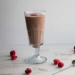 Side view- Single, tall glass with smoothie, topped with raspberries. Raspberries on table around the glass.