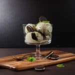Side view - Wooden cutting board with a glass dish filled with Mint Chocolate Cookie Ice Cream, topped with cookies.