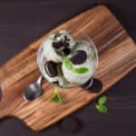 Wooden cutting board with a glass dish filled with Mint Chocolate Cookie Ice Cream, topped with cookies.