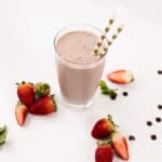 Tall glass filled with a Strawberry Mint Smoothie. Surrounded by strawberries and chocolate pieces.