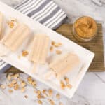 White tray with four Peanut Butter Pops, surrounded by peanuts and a jar of peanut butter.