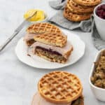 Prepared food image featuring waffle sandwiches with turkey, cranberry sauce, and stuffing. Side dishes include a white bowl of butter, a white bowl of red cranberry sauce, and a white dish of brown stuffing. Multiple golden waffles are visible. All items are on a white marble surface with a grey napkin.