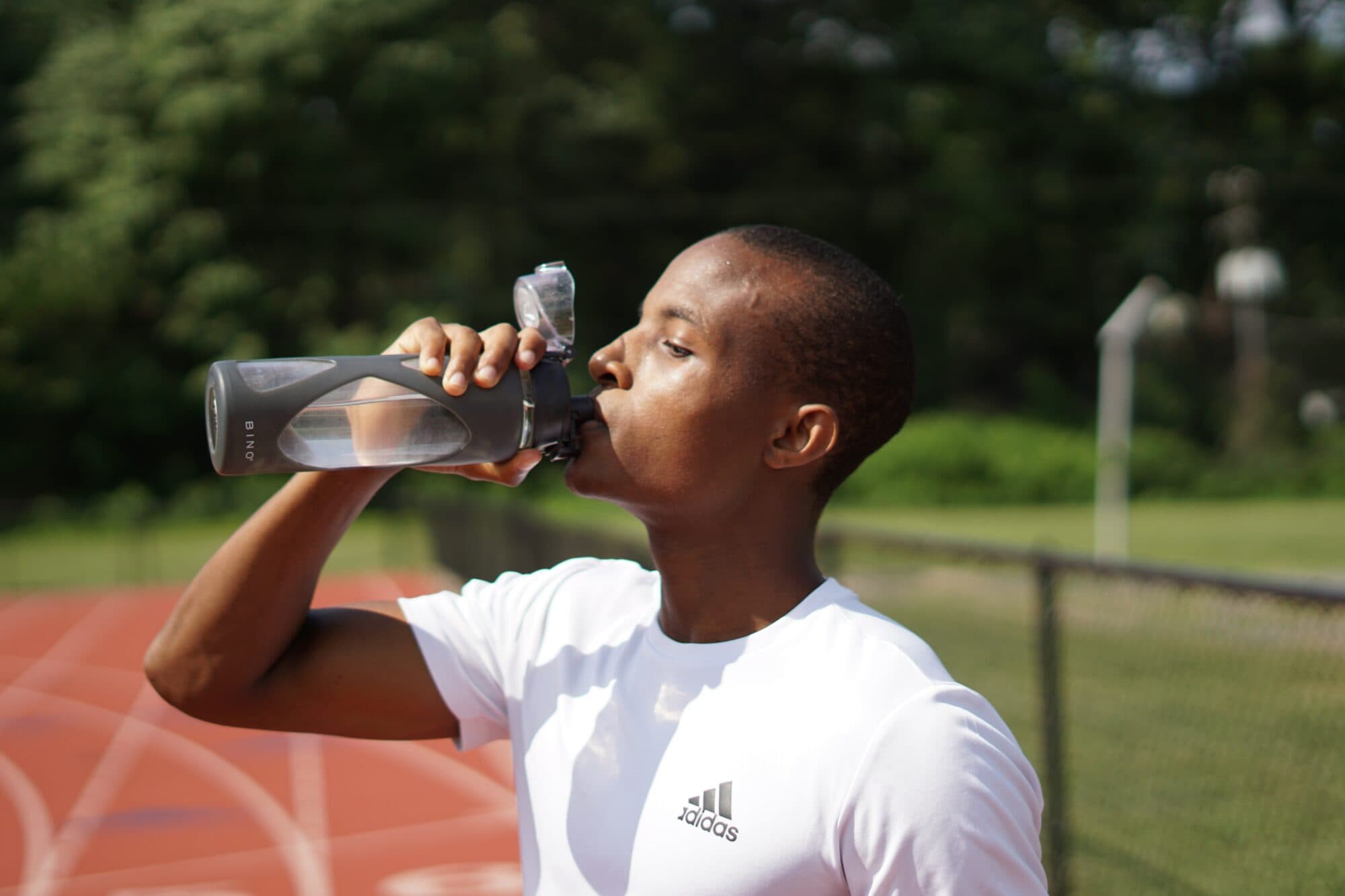Dark grey BINO brand reusable water bottle with clear sections, held by a person drinking. Person wears a white t-shirt with a dark grey adidas logo.