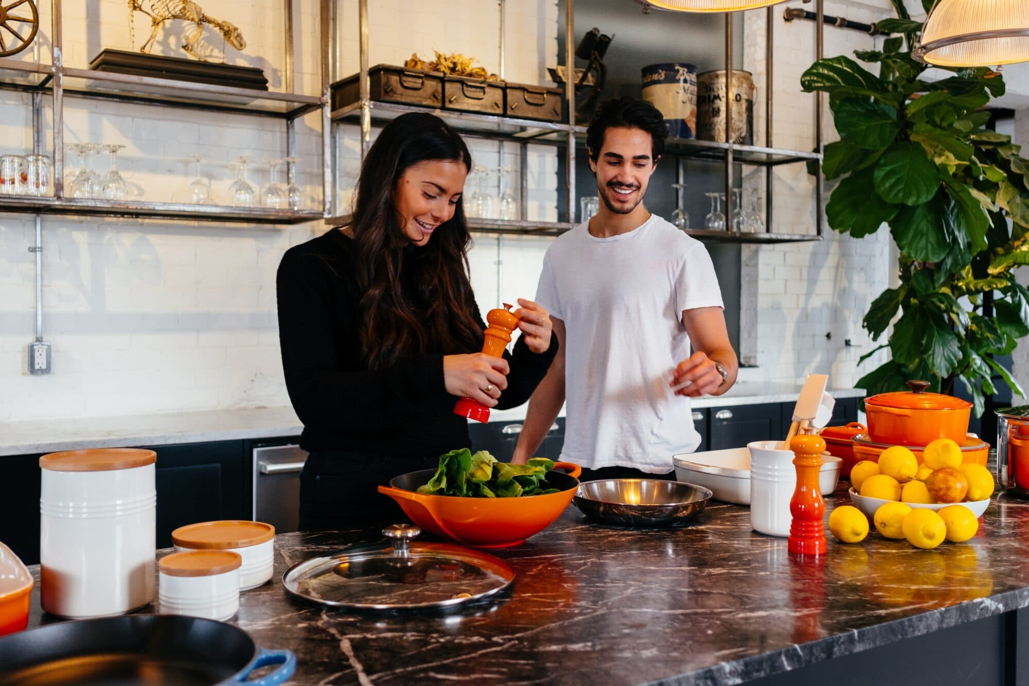 A man and a woman prepare food in a kitchen featuring various kitchenware. Visible products include three white LE CREUSET ceramic canisters with wooden lids, an orange enameled cast iron Dutch oven, an orange enameled cast iron shallow braiser, a black cast iron skillet with a blue handle, a red and orange salt and pepper mill, and a white ceramic utensil crock.