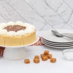 A round light brown cake with a dark brown top layer sprinkled with white salt flakes, encircled by white whipped cream, rests on a white cake stand. Multiple square caramel candies and a small clear glass bowl of white salt are scattered nearby. A stack of white plates with forks and an orange and white patterned napkin are also present on a white marble surface with a matching wall background.