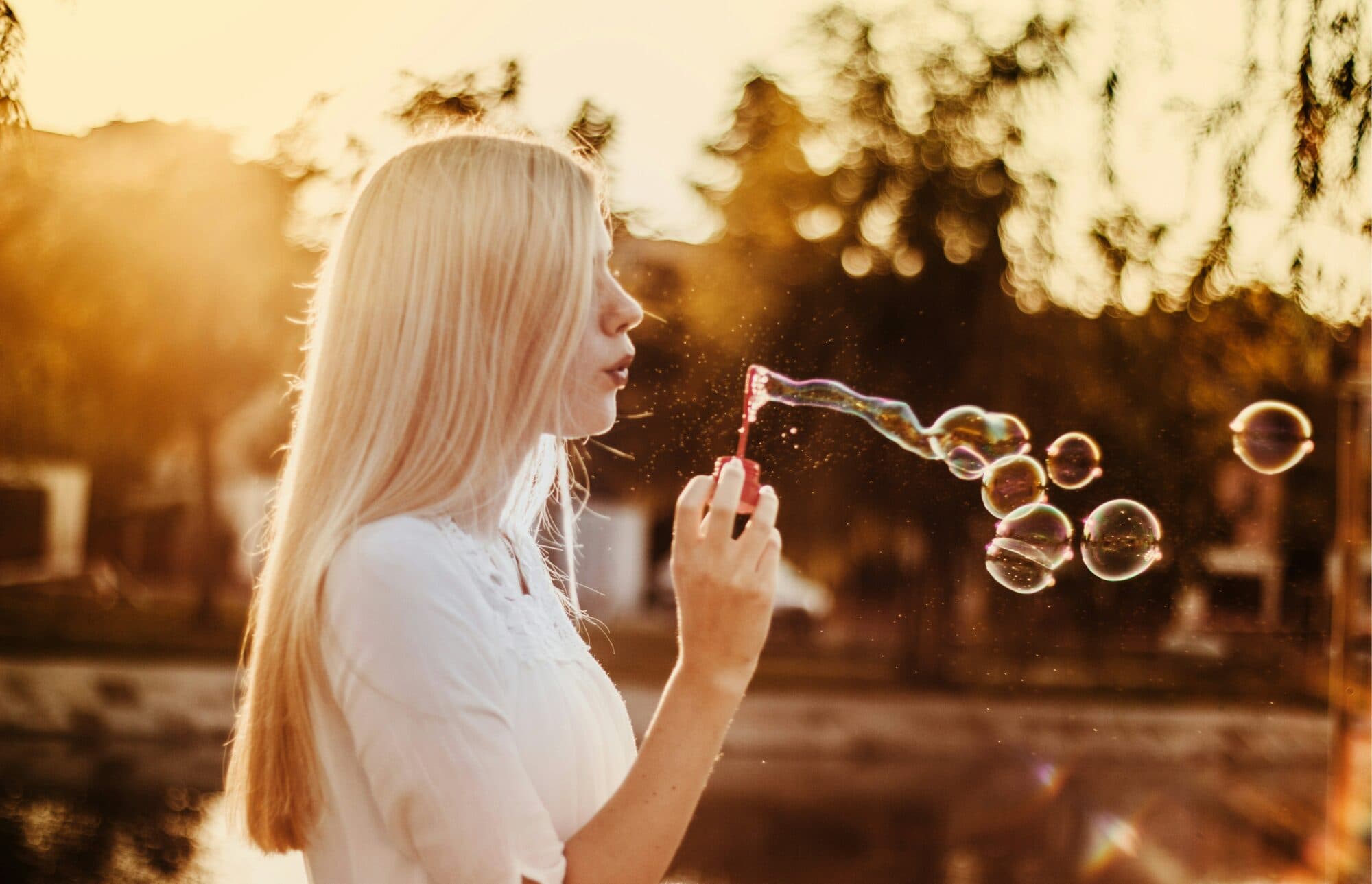 Young woman with blonde hair blowing bubbles from a red wand at sunset, with numerous iridescent bubbles floating in golden light.