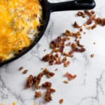 A baked casserole in a black cast iron skillet, topped with melted orange and white cheese. Cooked bacon pieces and shredded orange cheese are scattered on a white marble surface.